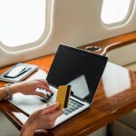 A woman holding a credit card and looking at her laptop while sitting in a fancy airplane seat.