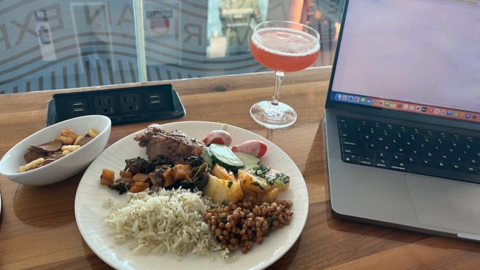 A plate of food, dish of snack mix and a cocktail on a table next to an open laptop with the American Express Centurion Lounge logo reflected in the glass behind.