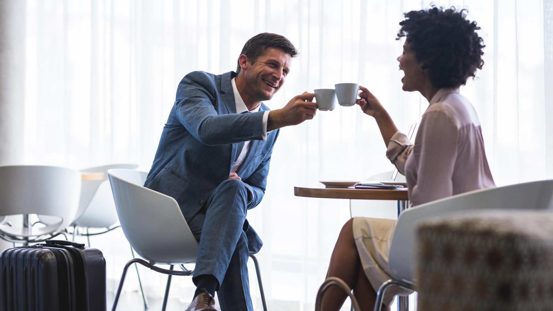 A man and women clink their coffee cups while sitting at a table in an airport lounge