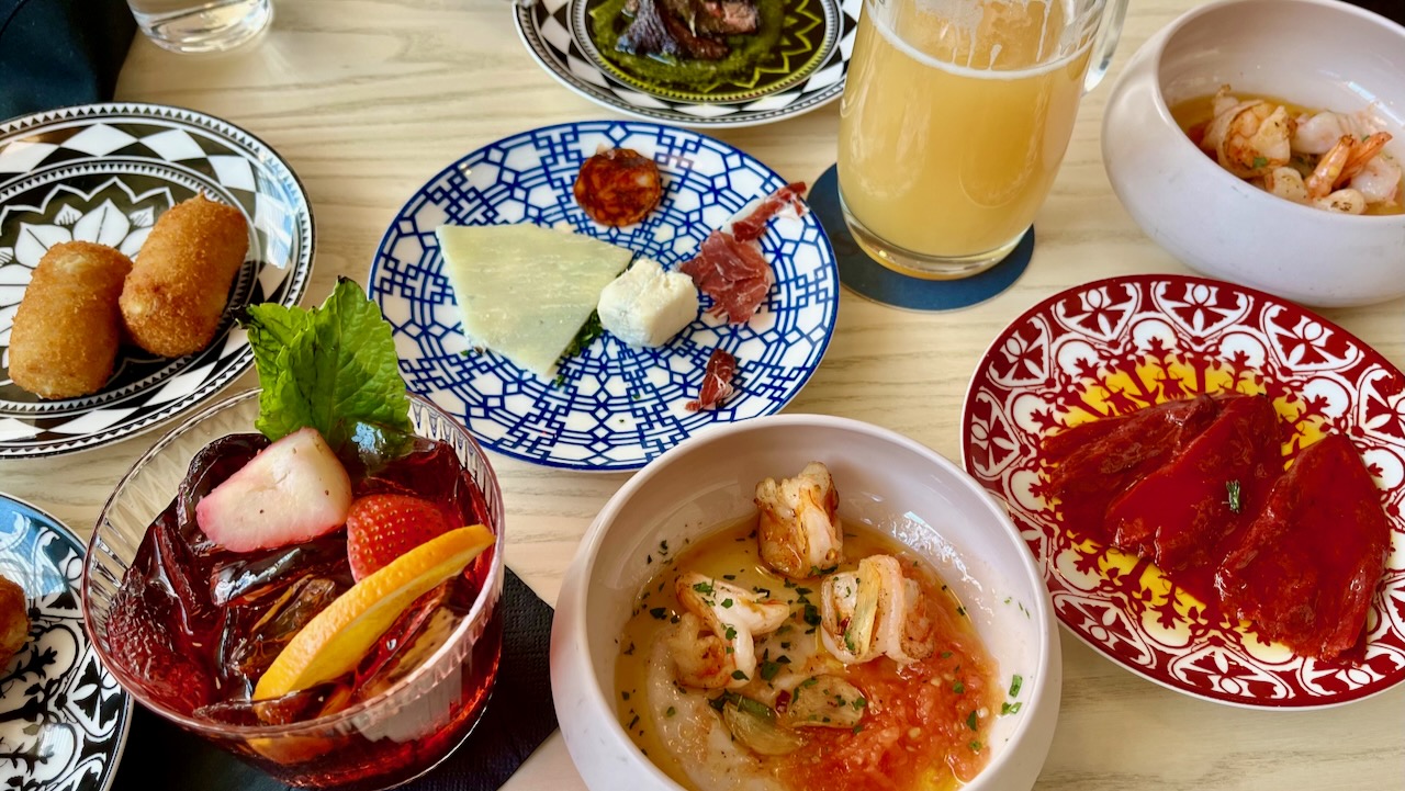 A table of various colorful tapas dishes including garlic shrimp, roasted peppers, sausage, croquettes and cheeses, along with a sangria cocktail and glass of beer.