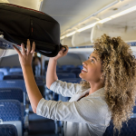 A smiling young businesswoman stands in the aisle of a commercial airliner and places her carry on bag in the overhead bin.