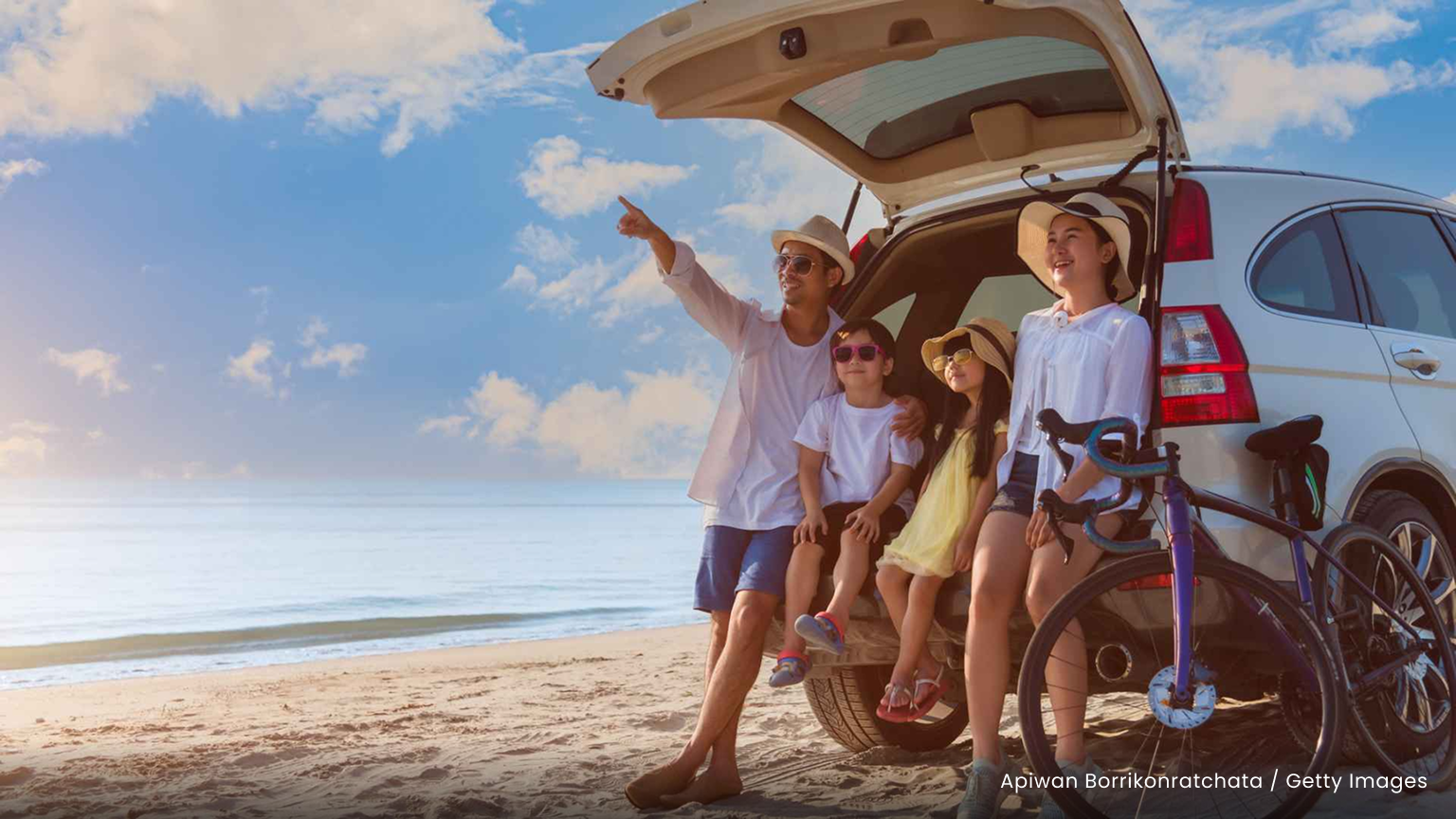 A father with three smiling young children points at something in the distance while they sit on the tailgate of their SUV parked on the beach.