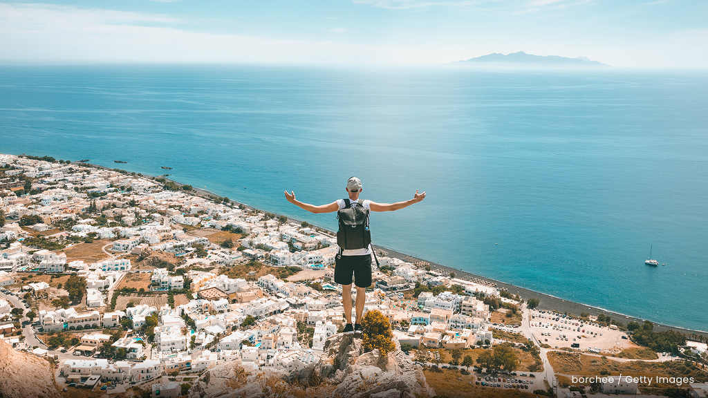 A man with a backpack with his arms spread wide stands on a cliff in Santorini overlooking a town and the ocean.