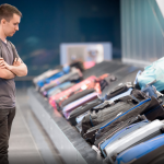 A pensive young man stands with his arms folded next to a baggage carousel filled with luggage.