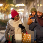 A couple walks down a street decorated with holiday lights; the man carries a small boy while the woman carries holiday shopping and packages.