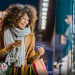 A young woman holding a credit card and holiday shopping bags stands outside a shop with a Christmas tree in the window.