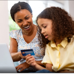 A mother and young daughter shop online with their laptop while the daughter holds a credit card.